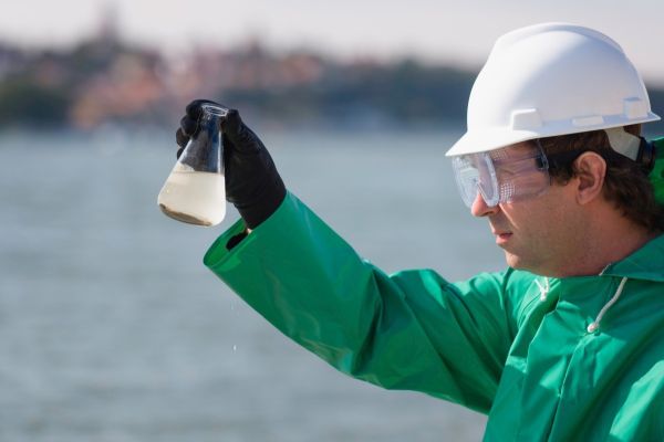 Umweltinspektor, mit Schutzbrille und weissem Schutzhelm, der eine Wasserprobe in einem Erlenmeyerkolben/Glas am Entnahmeort visuell überprüft