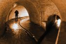 Ein Kanalarbeiter in Gummistiefeln und mit Stirnlampe reinigt mit einer Schaufel eine Rinne in der Kanalisation in der Nähe einer Einmündung eines kleineren Kanals. Eine Stromkabelrolle steht an der linken Wand und ist teilweise abgerollt.