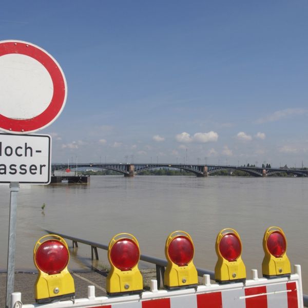 Ein Hochwasser-Warnschild und eine Absperrbake weißt auf Hochwasser am Rhein hin. Eine Brücke führt über den Rhein bei Mainz. Ein Segelschiff liegt am gegenüberliegenden Ufer vor Anker.