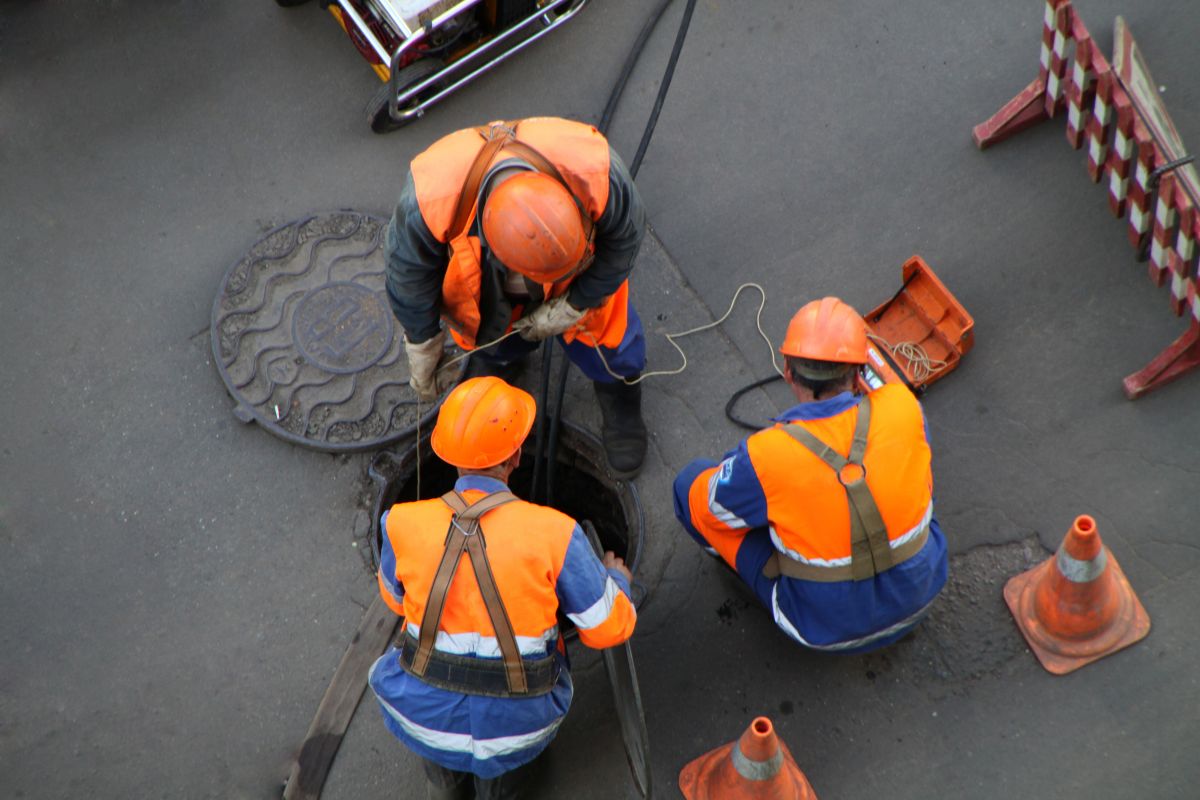 Drei Arbeiter mit Schutzhelmen an geöffnetem Kanaldeckel