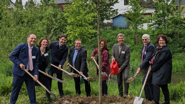 Ein neuer Baum wird eingepflanzt und verschiedene Personen, die zum Jubiläum eingeladen waren, sind mit Spaten und Gießkanne rund um einen Baum zu sehen