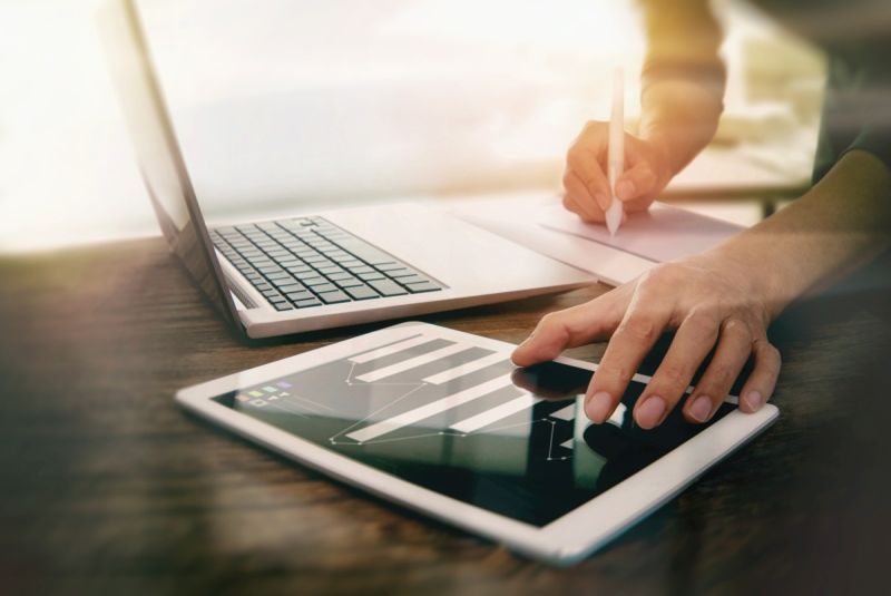 Vor einem geöffneten Laptop steht eine Person und notiert et- was mit der rechten Hand. Mit der anderen Hand bedient sie ein Tablet, auf dem ein geöffnetes Programm mit einem Säulendiagramm zu sehen ist.