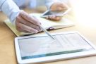 Eine Person prüft eine Abrechnung auf einem Tablet-Computer. Es ist eine Nahaufnahme der Hände der Person zu sehen, die einen Kuli und ein Handy halten. Ein Notizbuch ist aufgeschlagen. Davor liegt das Tablet.