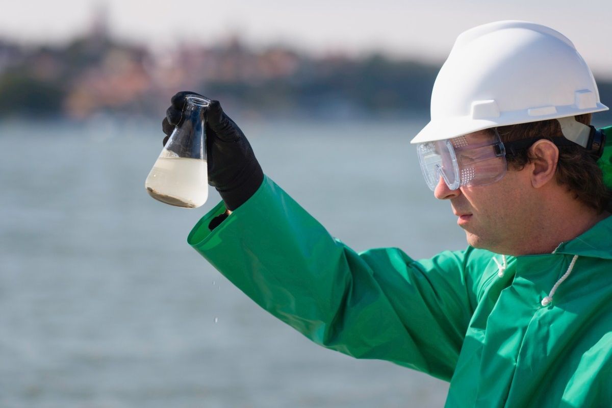 Umweltinspektor, mit Schutzbrille und weissem Schutzhelm, der eine Wasserprobe in einem Erlenmeyerkolben/Glas am Entnahmeort visuell überprüft