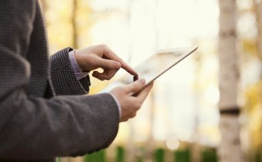 Ein Mann im karierten Anzug tippt mit dem linken Zeigefinger auf ein Tablet, das er in der rechten Hand hält.