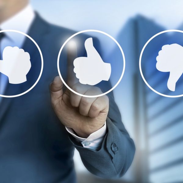 Hinter einer Glasscheibe mit 3 Emojis Buttons mit Daumen hoch, runter und nach links steht ein Geschäftsmann, der mit dem linken Zeigefinger auf den mittleren Button mit Daumen nach oben zeigt.