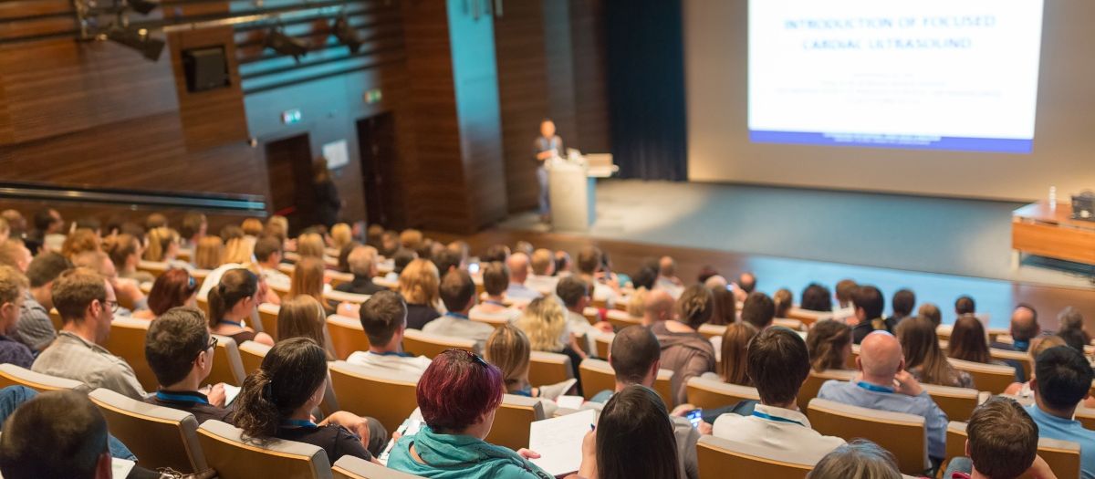 ein gut gefüllter Hörsaal, in dem viele Menschen einem Vortrag auf einer großen Leinwand folgen