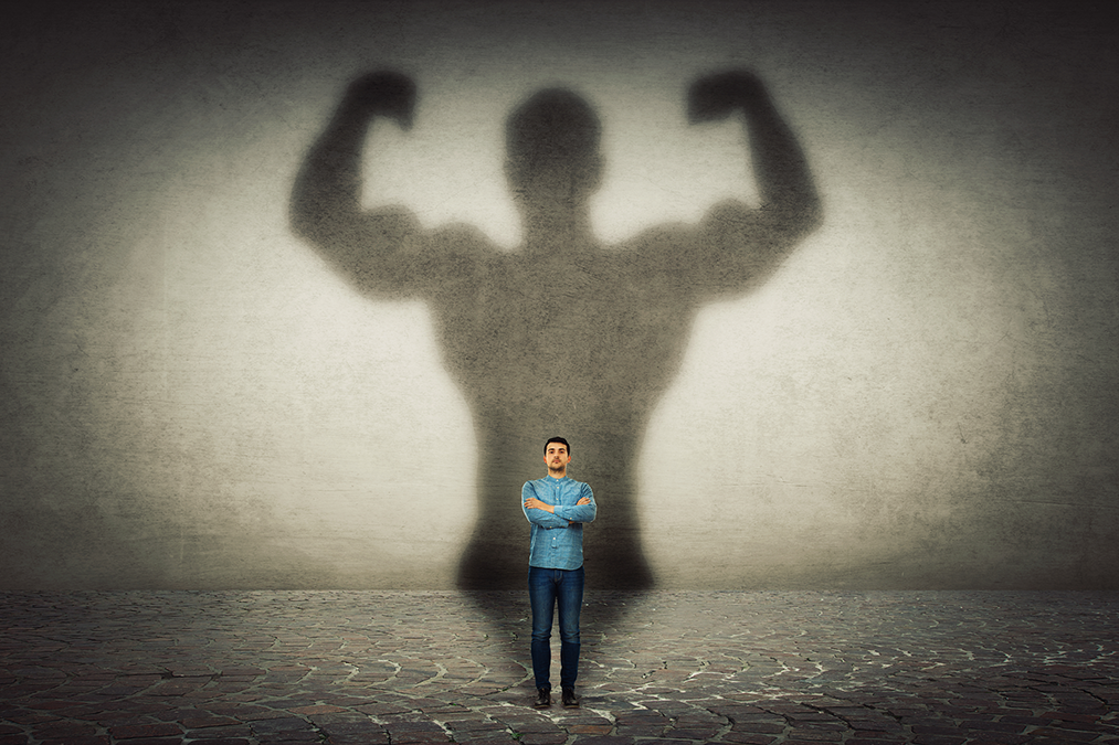 Mann mit verkreuzten Armen steht vor seinem Schatten mit starken Muskelarmen