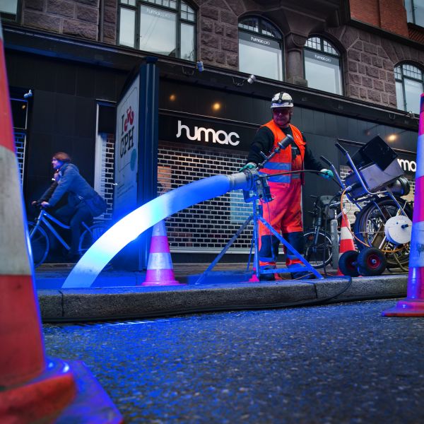 Ein Kanalarbeiter mit Helm und Schutzkleidung bedient einen LED-Strahler für die Lichthärtung eines Schlauchliners auf einer Baustelle. Die Baustelle, die sich an einer Geschäftsstraße befindet, ist mit Pylonen und Baustellenschild abgesichert. Eine Radfa