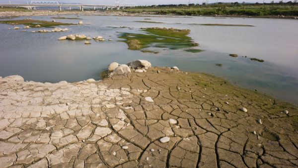 Fluss führt wenig Wasser. Teilweise mit rissigen ausgetrockneten Stellen. Eine Brücke und Starkstrommasten im Hintergrund.