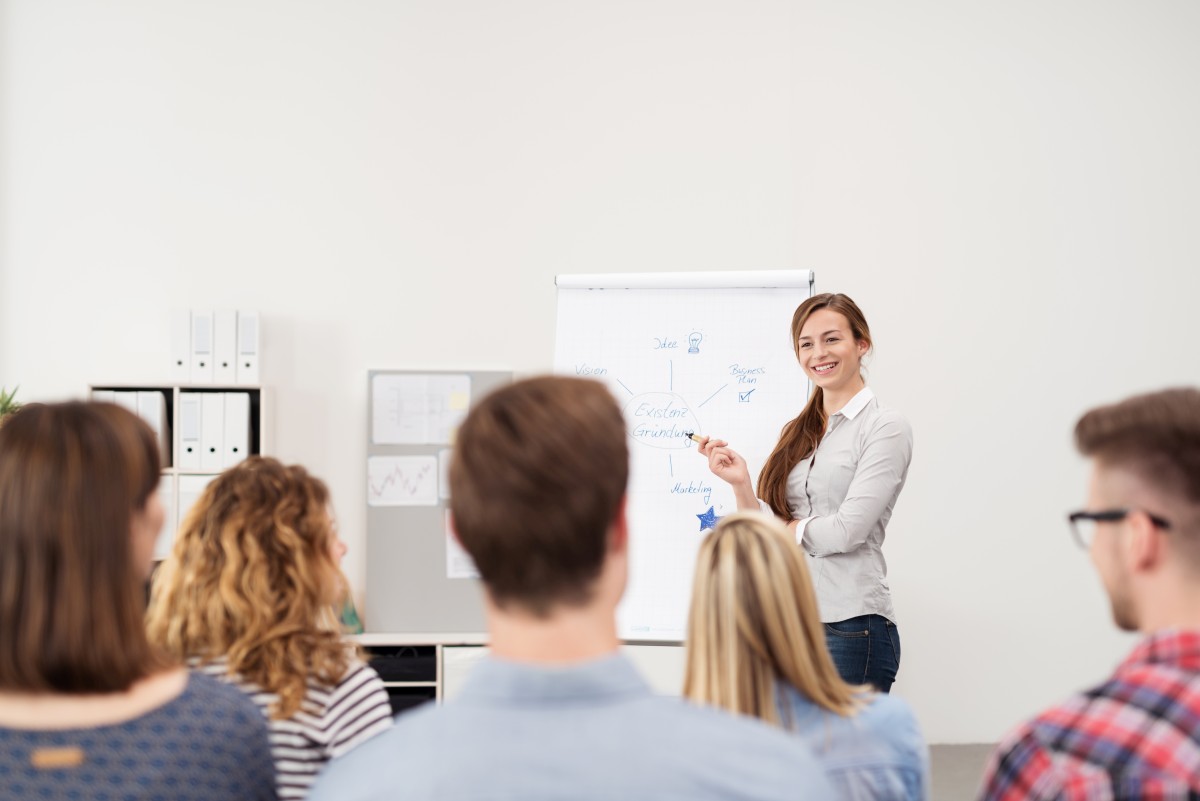 Junge Frau hält einen Vortrag vor Stundenten über Existenzgründung mit Hilfe einer Grafik am Flipchart.